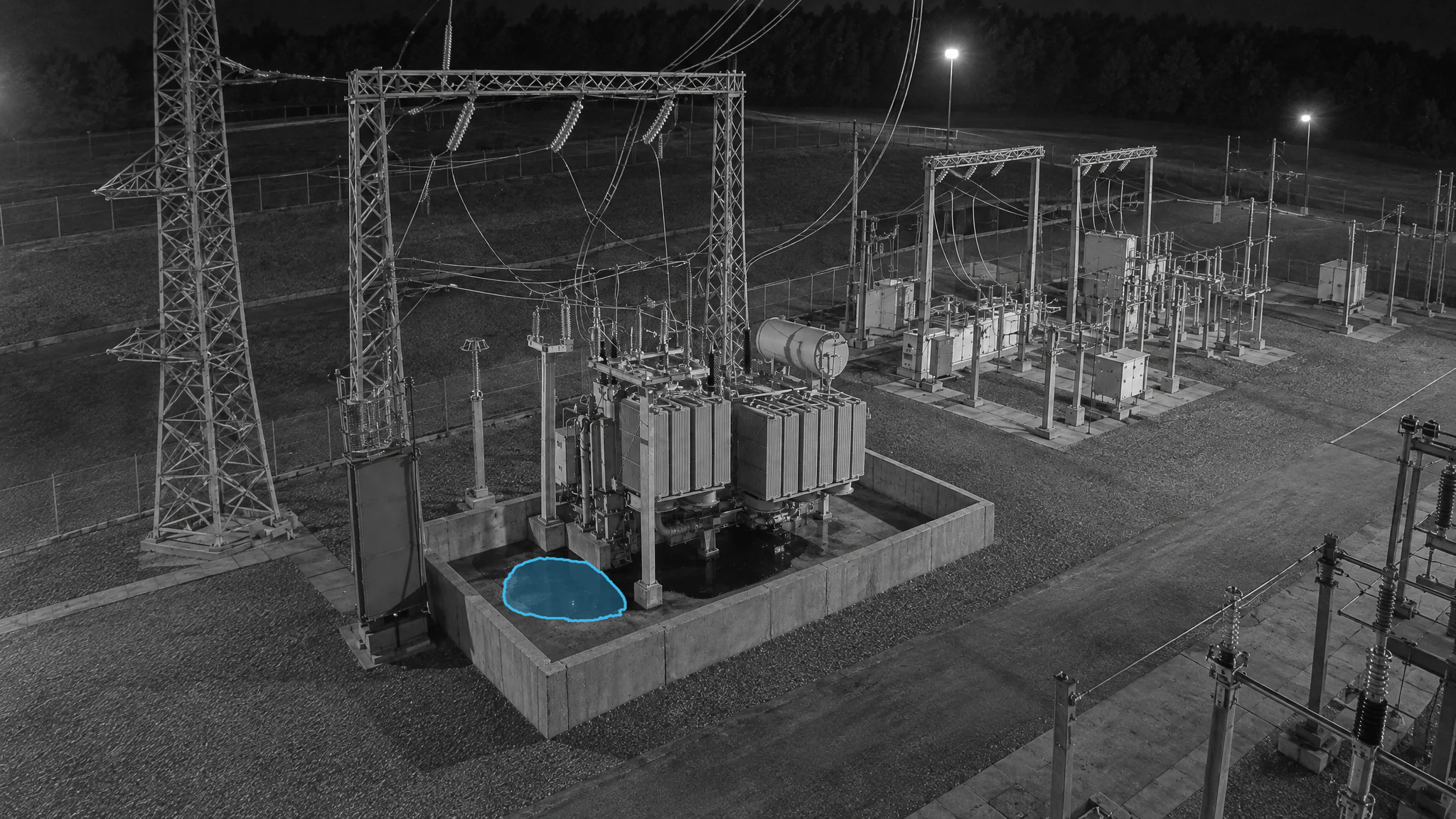 Monochrome nighttime view of a transformer inside concrete containment, with a blue puddle overlay in the corner.
